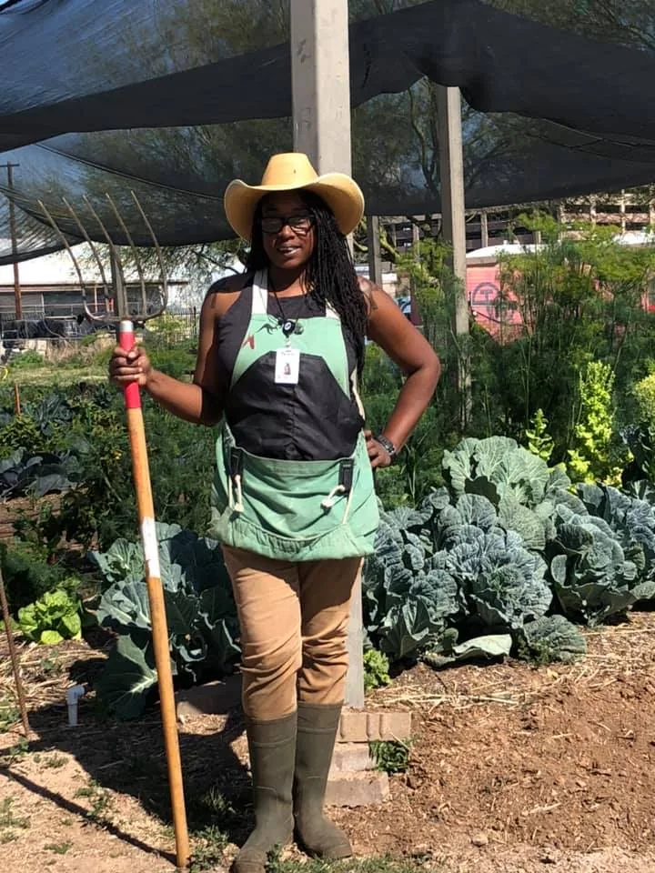 Nika Forte' standing in a shaded garden bed holding a pitchfork, wearing a farm apron and hat